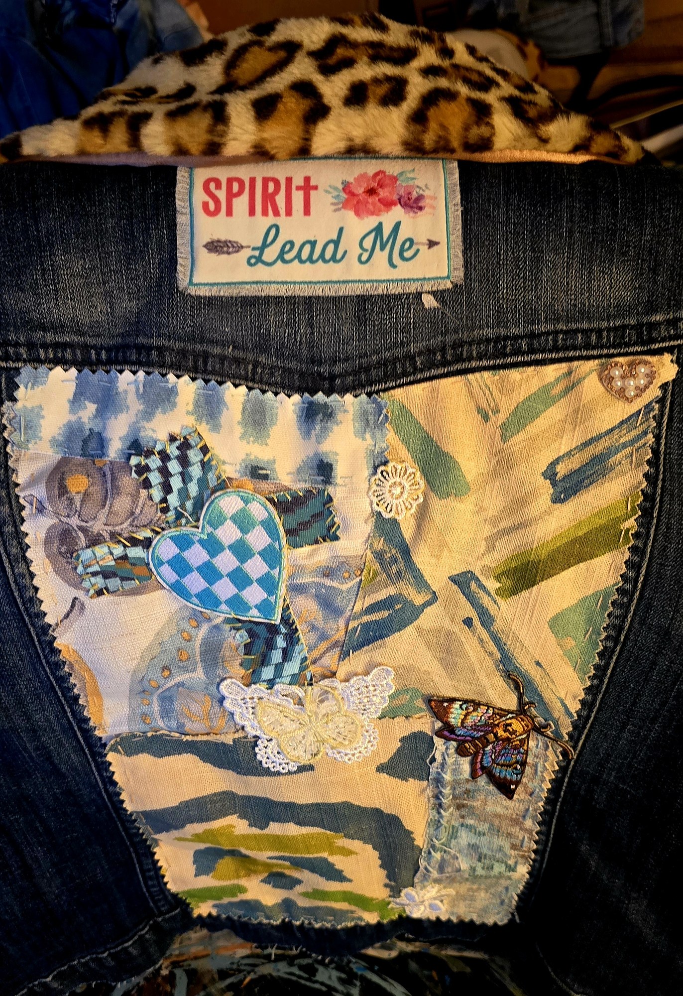 Close detail of a denim Memory Jacket with a Spirit Lead Me patch, heart appliqué, butterfly, and stitched fabrics