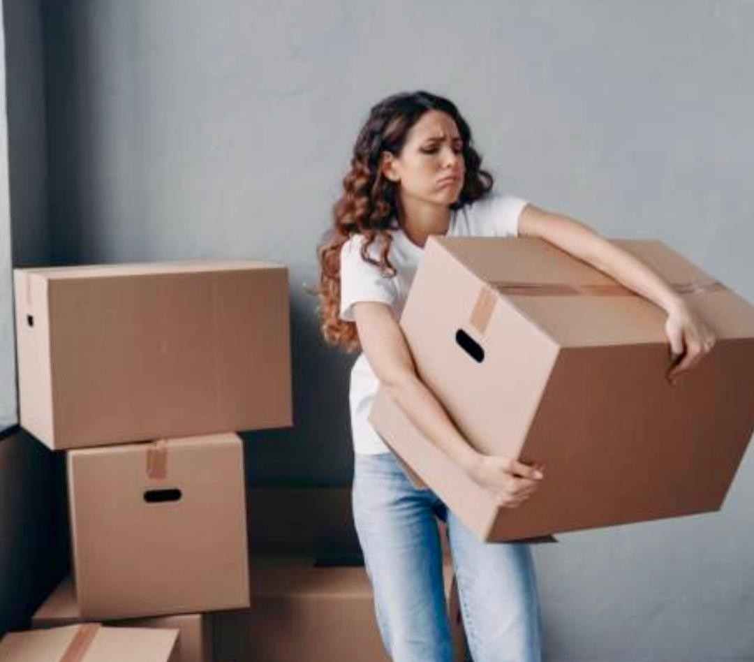 A woman carrying a large cardboard box beside stacked boxes