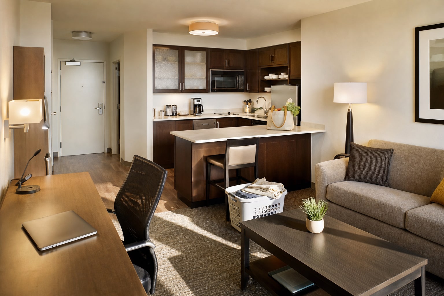 An extended-stay hotel suite arranged with a desk, kitchenette, laundry basket, and living area