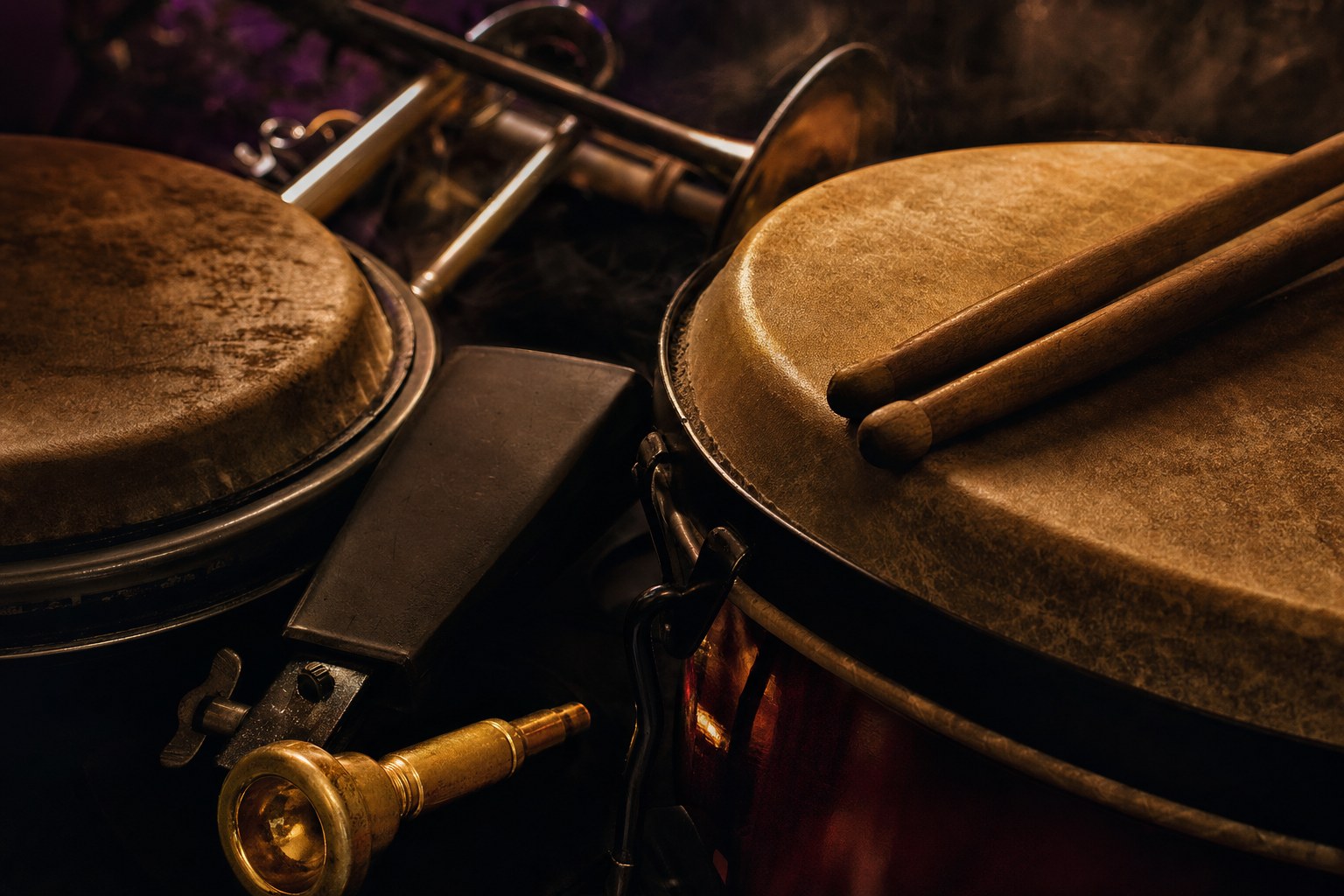 Hand drums, drumsticks, cowbell, and brass mouthpiece in low warm light