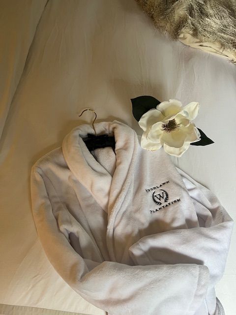 A white Woodlawn Plantation robe laid on a guest bed beside a magnolia flower