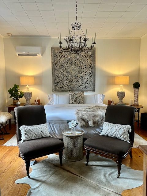 A guest bedroom with an ornate headboard, chandelier, and armchairs